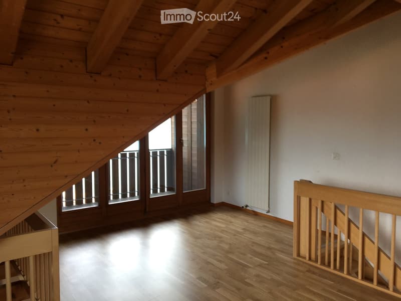 Empty attic space with wooden beams, wooden floor, and large windows with metal bars, featuring a wooden railing and radiator on the wall.