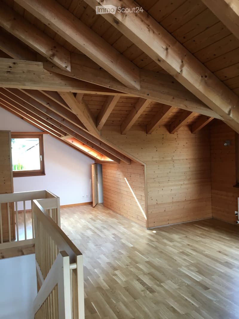 An empty attic space with exposed wooden beams, wooden flooring, and a small window letting in natural light. The room features a white railing and a partially visible staircase leading downwards.
