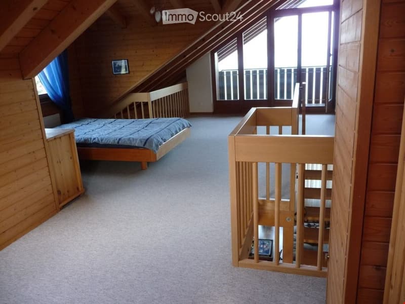 Spacious attic bedroom with wooden beams, large windows, and a raised platform area. Features a single bed, nightstand, and a small picture frame on the wall.