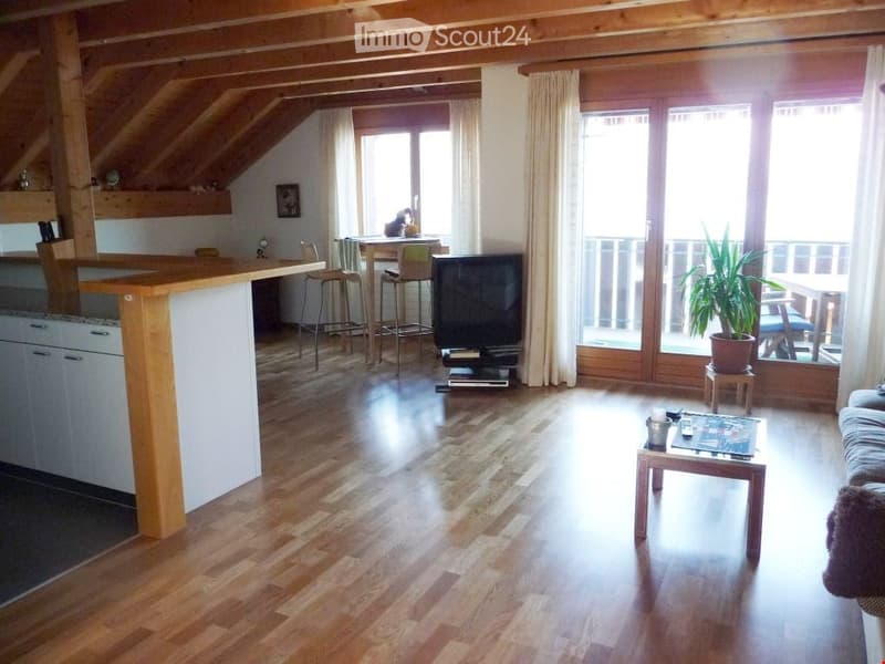 Open-plan living and dining area with wooden floors, exposed wooden beams, large windows, and a balcony. Features a sofa, coffee table, television, dining table with chairs, and a kitchen island.