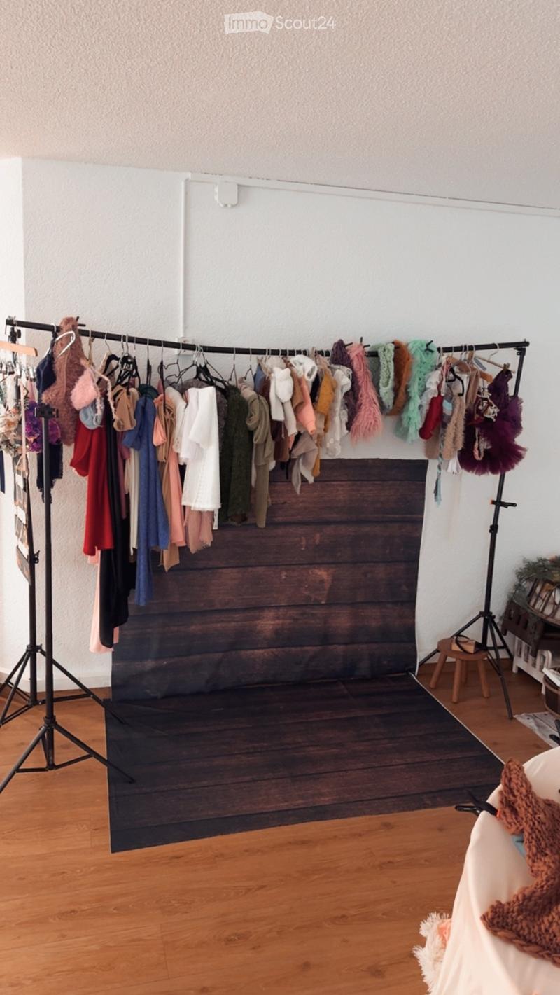 A room with wooden flooring and a large backdrop, featuring a clothing rack with various garments, a small stool, and a table with a blanket draped over it.