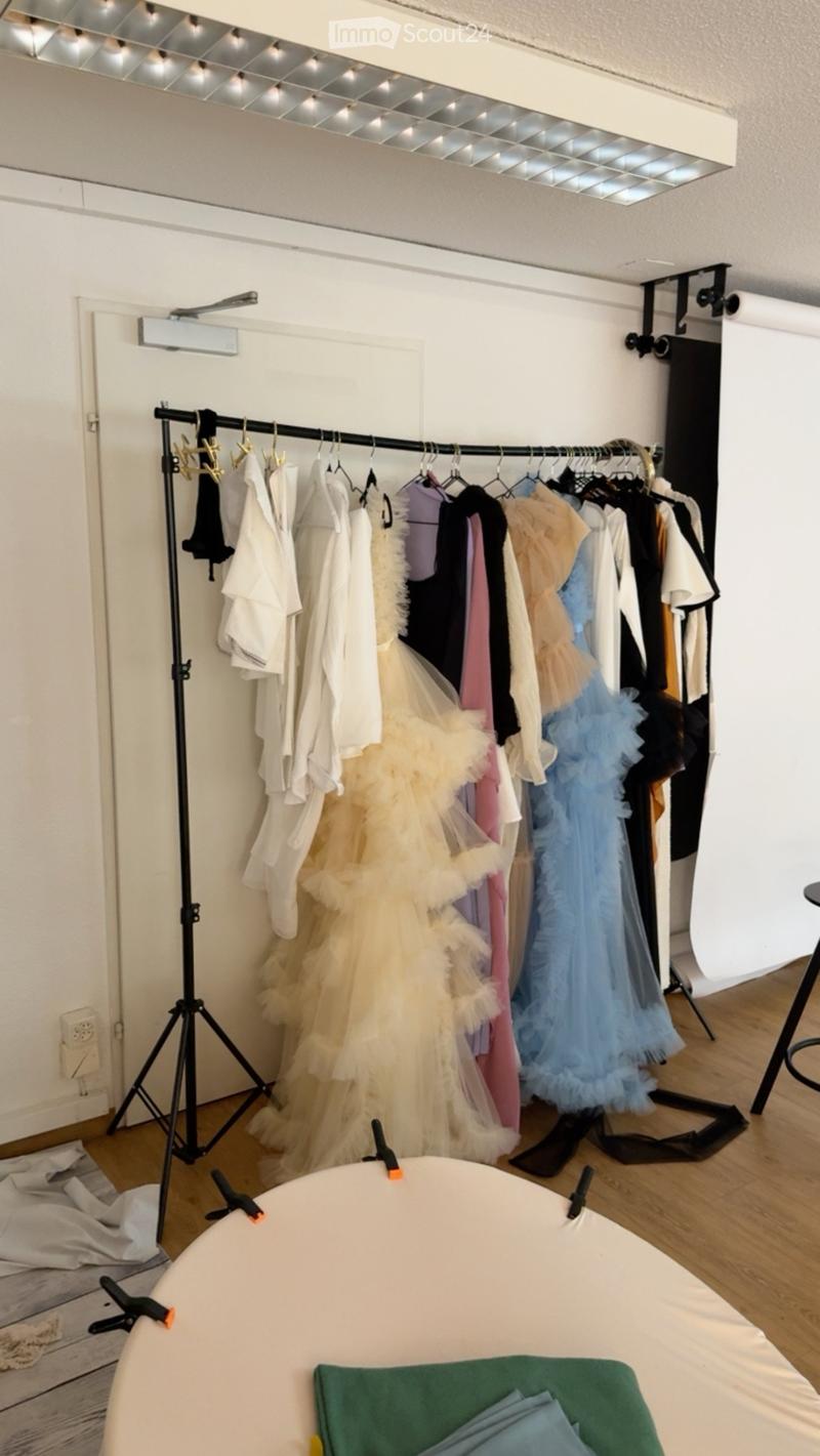 A well-lit room with a clothing rack holding various dresses, a tripod, a stool, and a table with folded fabrics.