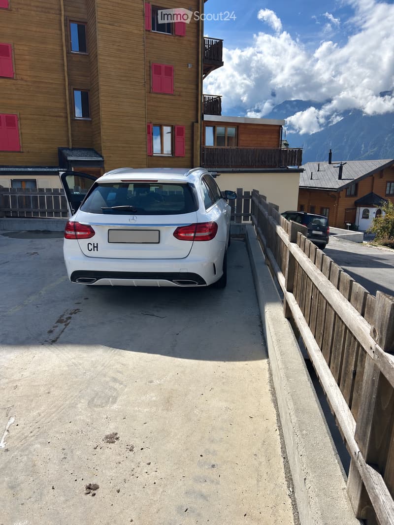 A parking area with a white Mercedes-Benz car, wooden fence, and a multi-story wooden building with red shutters and balconies in the background.