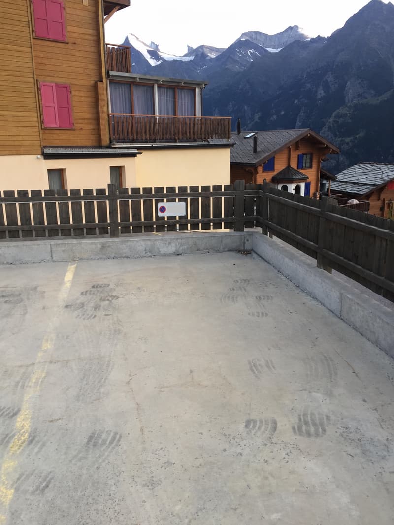 Concrete balcony with wooden railing, overlooking a mountainous landscape with snow-capped peaks.