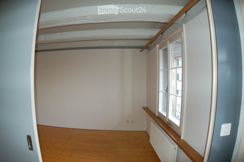 Empty room with wooden flooring, white walls, and a window with white frames. The room features a radiator under the window and exposed wooden beams on the ceiling.