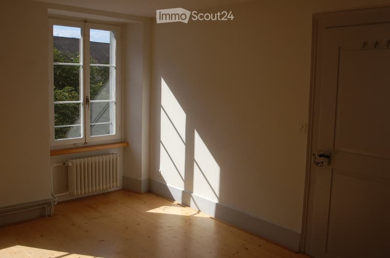 Empty room with wooden flooring, white walls, a window with a radiator below, and a closed door.