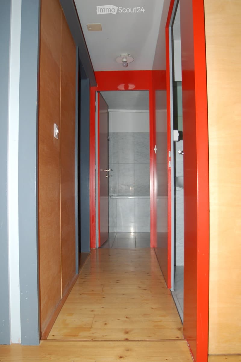 Wooden floor hallway with red and grey walls, featuring a light fixture and a partially visible bathroom at the end.