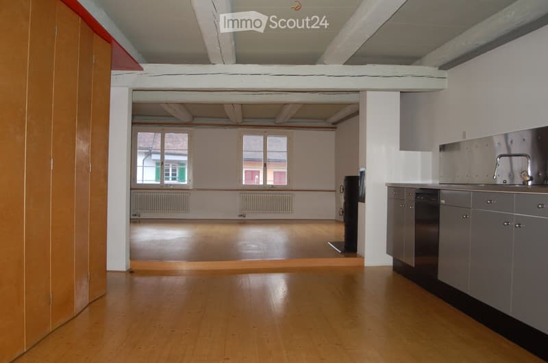 An unfurnished room with wooden flooring, white walls, and exposed wooden beams on the ceiling. A kitchen area is visible with cabinets and a sink, but no furniture or appliances are present.