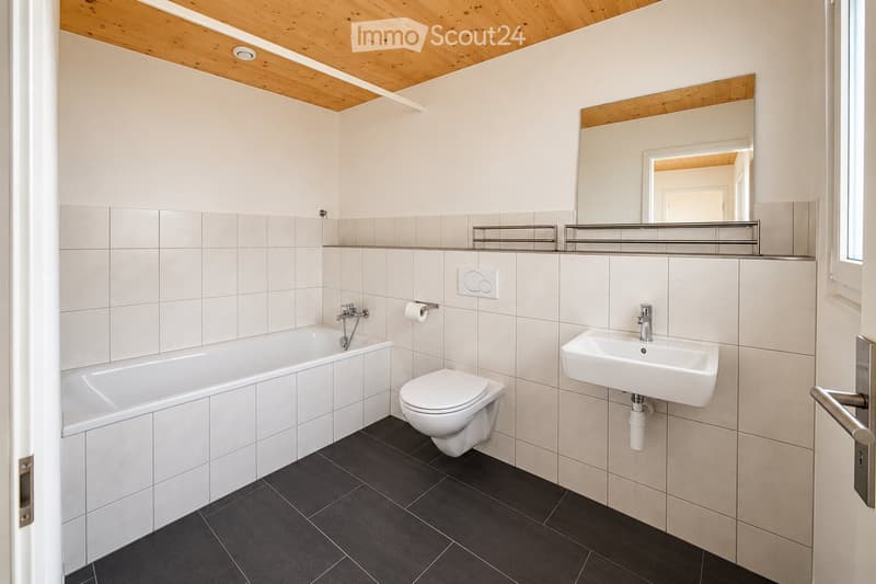 Modern bathroom with white tiles, a bathtub, toilet, sink, and a large mirror. Features a wooden ceiling and dark grey floor tiles.
