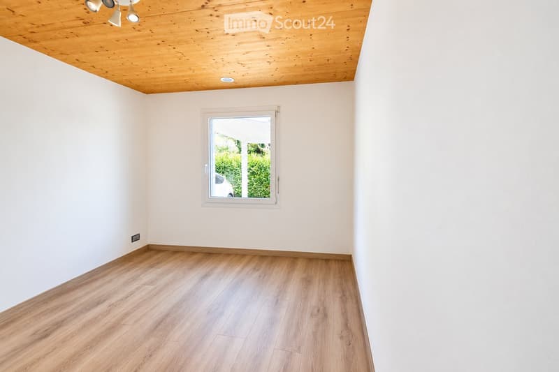 Empty room with light wooden flooring, white walls, and a large window with a view of greenery outside.