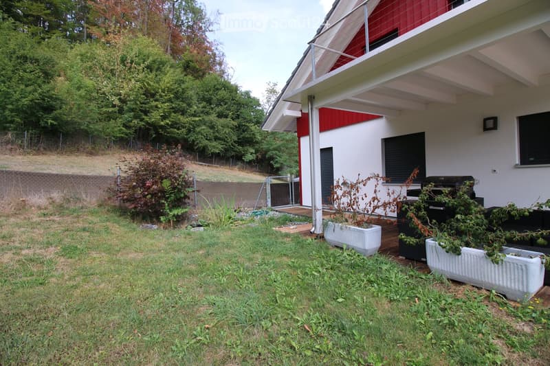 A modern two-story house with white and red exterior, featuring a covered patio area with potted plants and a barbecue grill. The surrounding yard is grassy with a fence and trees in the background.