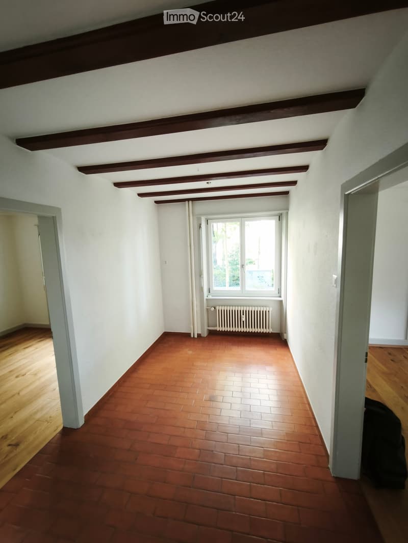 Empty room with wooden flooring, white walls, exposed wooden beams on the ceiling, and a window with a radiator below it.
