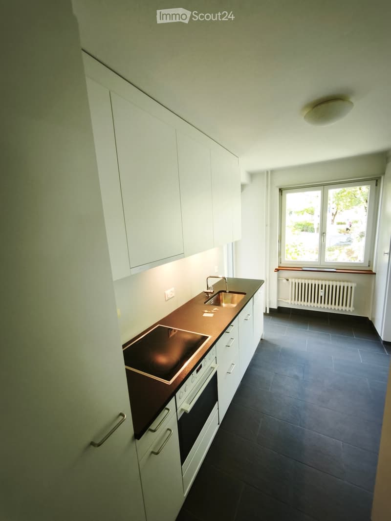 Modern kitchen with white cabinets, dark countertops, built-in oven, sink, and a window overlooking a green area.