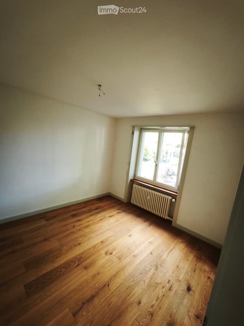 Empty room with wooden flooring, white walls, and a single window with a radiator below it.