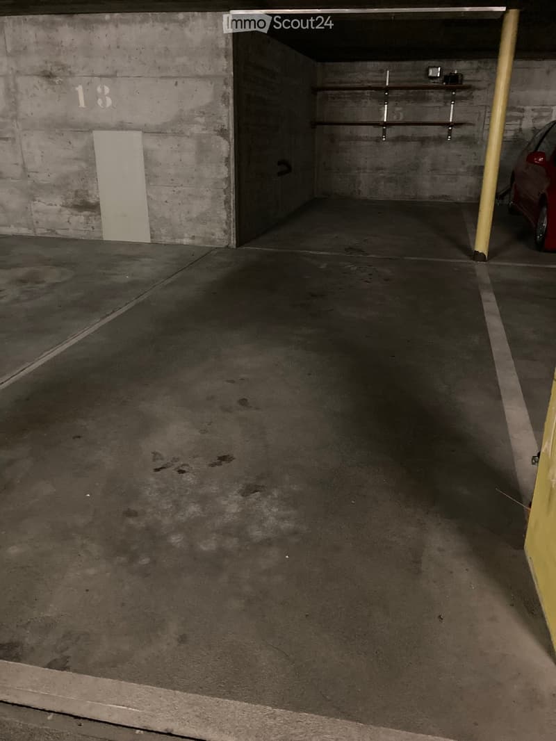 Empty underground parking space with concrete floors and walls, marked with number 13, featuring a single parking spot and a small storage area with shelves.
