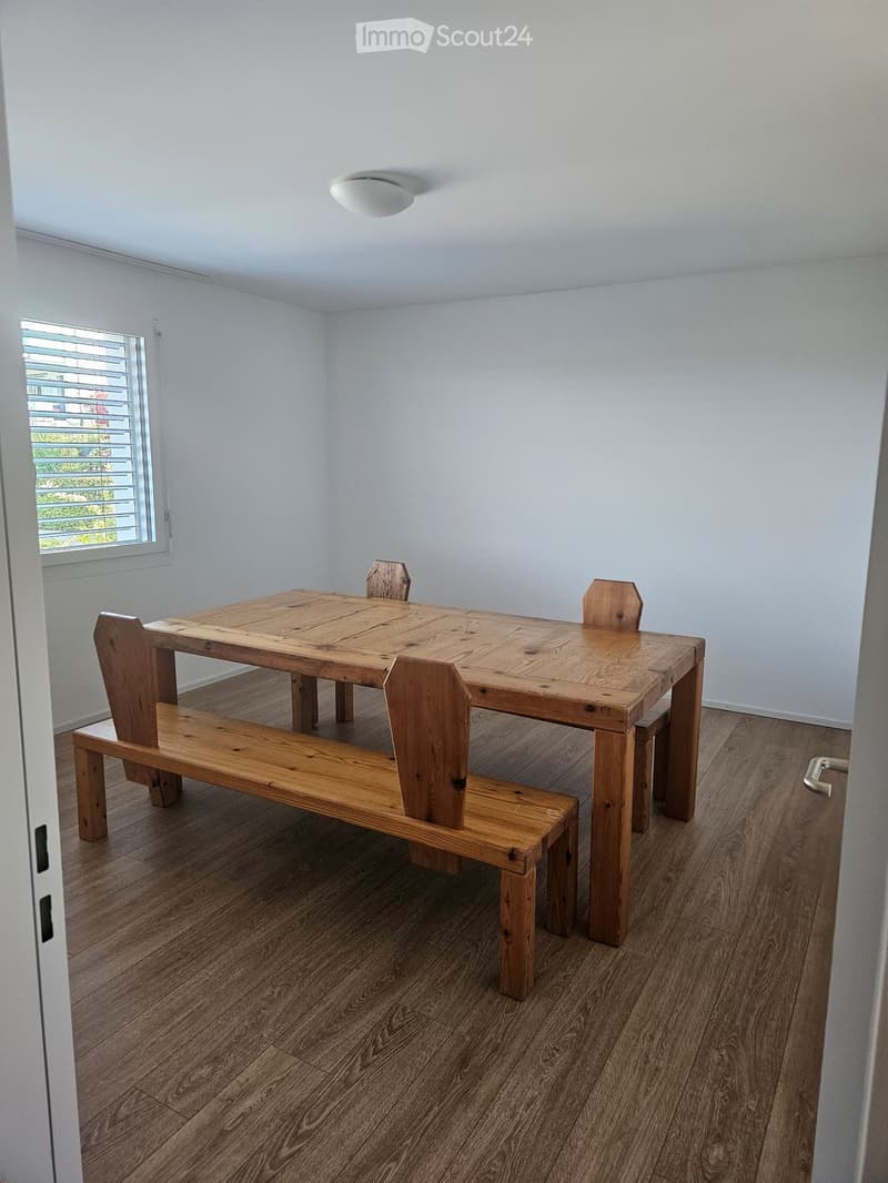 A room with a large wooden dining table, two benches, and four chairs. It has white walls, a wooden floor, and a white ceiling light.