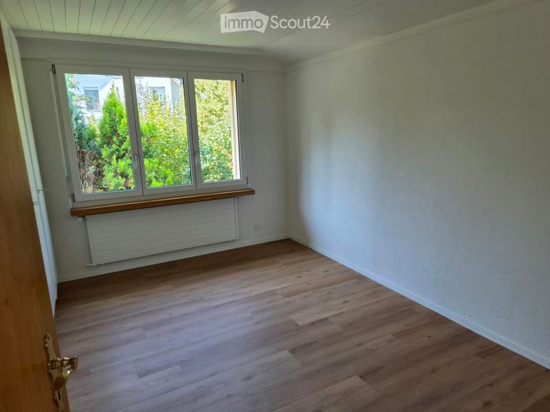 Empty room with wooden flooring, white walls, large windows, radiator, and a door.