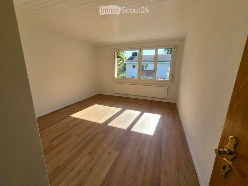 Empty room with wooden flooring, white walls, and a large window with a view of neighboring houses. A radiator is visible below the window.