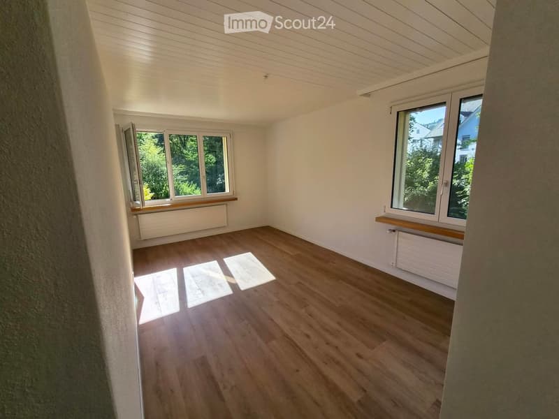 Empty room with wooden flooring, white walls, and large windows with wooden sills. Natural light streams in, casting shadows on the floor.