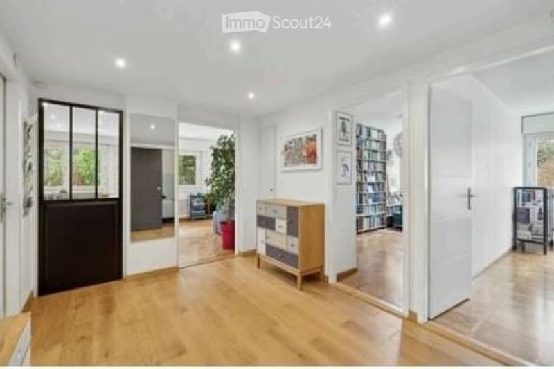 Bright, open-plan living space with light wooden flooring, white walls, and modern decor. Features include a storage unit, bookshelf, potted plant, and large mirror on the wall.