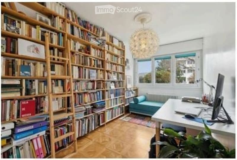 A cozy, well-lit study or home office with built-in wooden bookshelves filled with books, a large window offering a view, a comfortable blue sofa, a modern desk with a laptop and computer monitor, and a decorative chandelier.