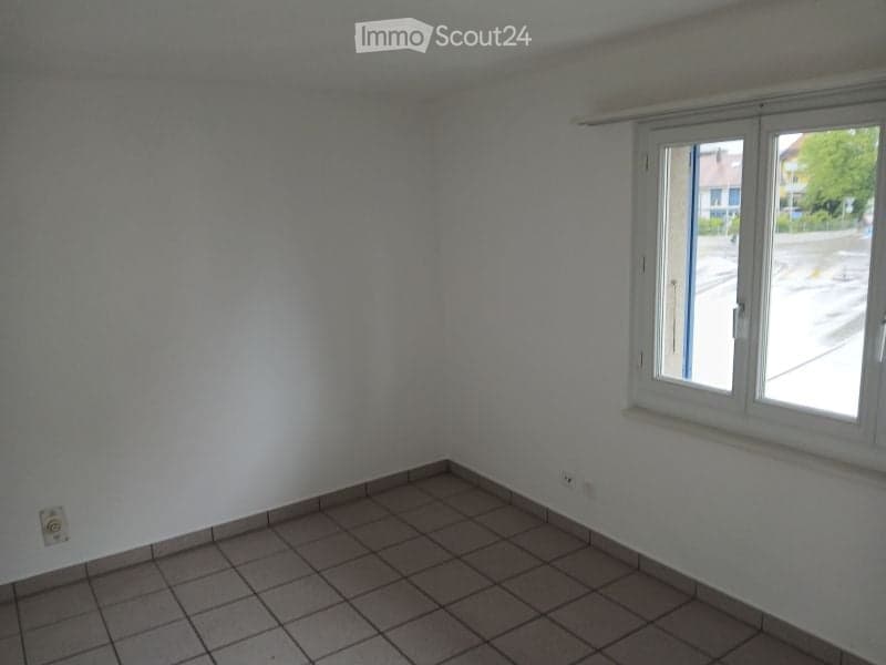Empty room with tiled floor and white walls, featuring a large window with white frame and a visible electrical outlet on the wall.