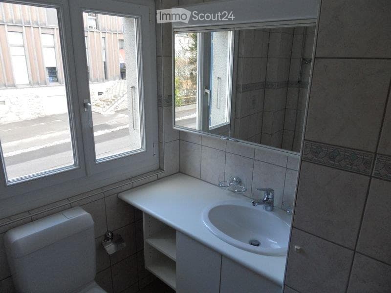 Modern bathroom with a white sink, chrome faucet, large mirror, and toilet. The room has tiled walls and a window offering a view of the street.