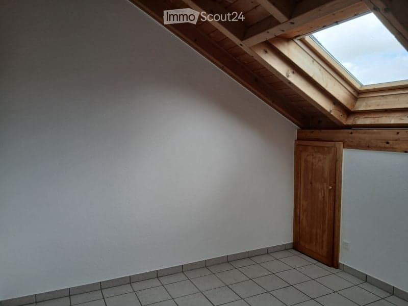 Empty attic room with wooden ceiling beams, skylight window, tiled floor, and a wooden door.