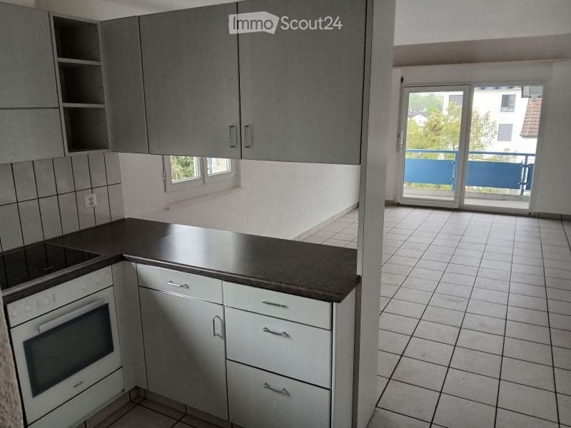 Modern kitchen with grey cabinets, white countertop, and an oven. Large windows and a sliding glass door lead to a balcony.