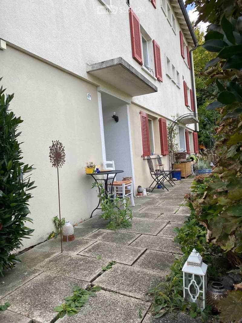 A small, paved outdoor area adjacent to a building with red shutters. Features include a small table and chairs, potted plants, and decorative items.