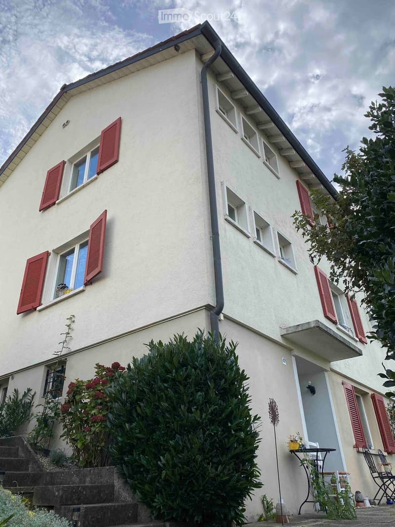 Two-story house with white exterior, red shutters, multiple windows, and a small balcony area with outdoor furniture.