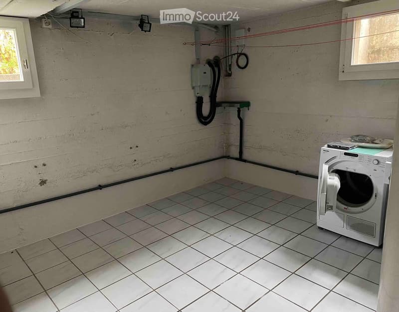 Small utility room with tiled floor, concrete walls, and a white washing machine. There are two windows, electrical outlets, and visible pipes.