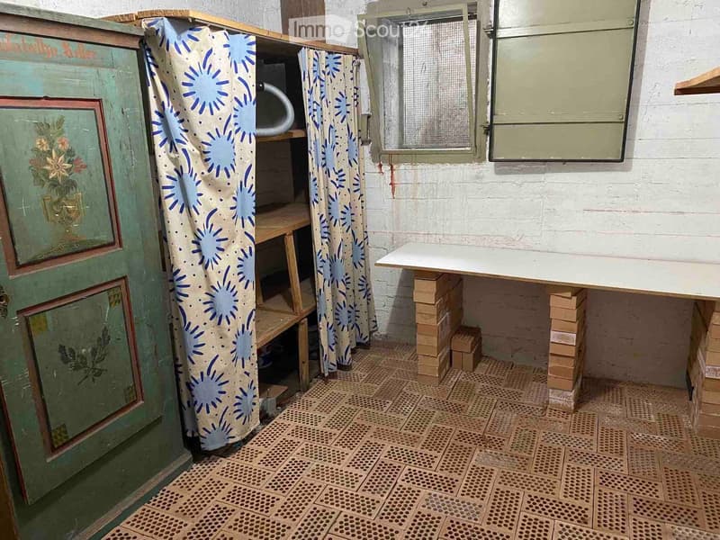 Empty room with exposed brick walls, tiled floor, and a wooden shelf partially covered by a blue and white curtain. A small table is supported by stacked bricks.