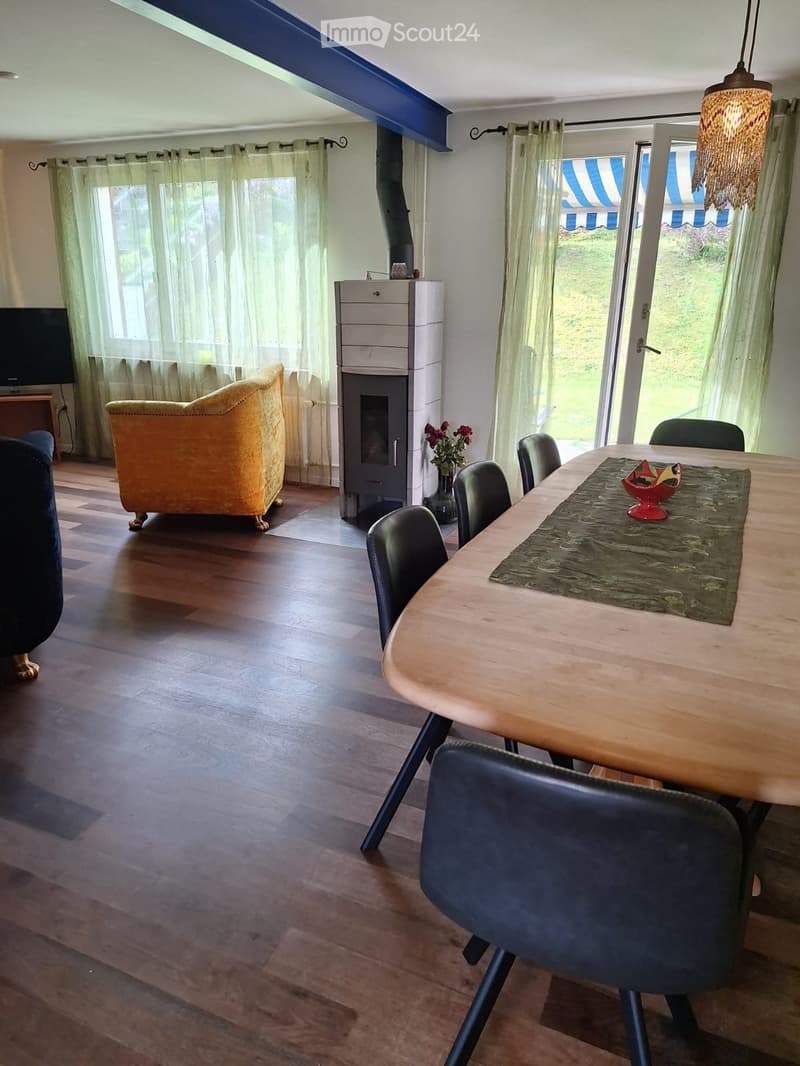 A cozy living and dining area with a yellow sofa, a wooden dining table with chairs, a modern fireplace, and large windows with curtains.