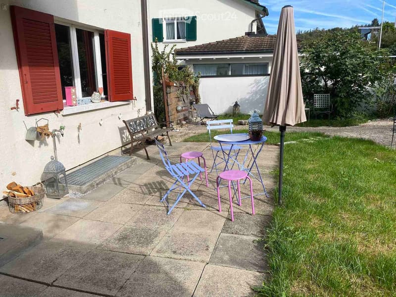 Outdoor patio area with a small table, chairs, umbrella, bench, lantern, and a basket of firewood near the house entrance.