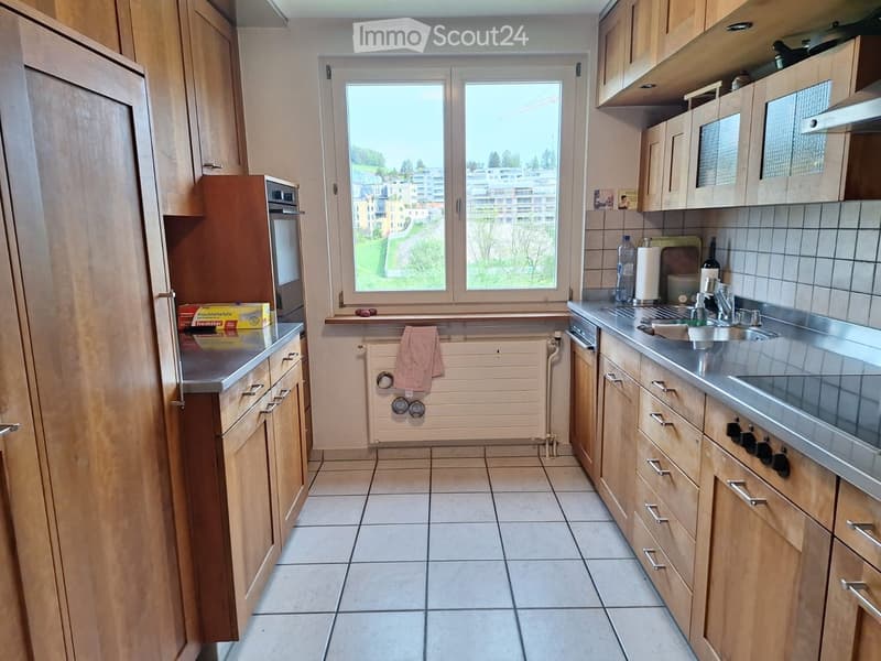 Modern kitchen with light wooden cabinets, stainless steel appliances, tiled floor, and a window overlooking a residential area.