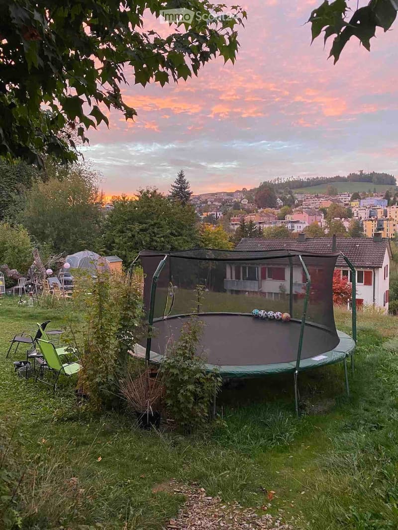 Garden with trampoline, chairs, table, and balls, overlooking a town with a sunset view.