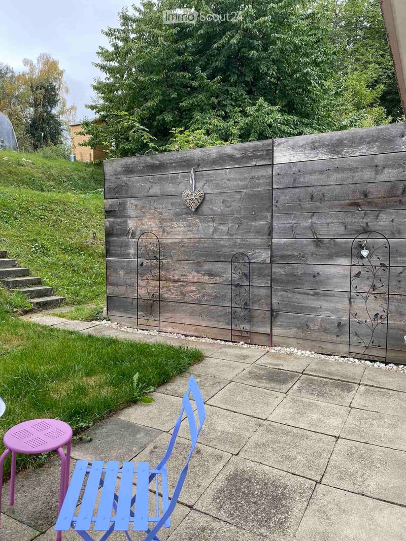 Garden area with a concrete patio, wooden fence, and two chairs, one pink and one blue.