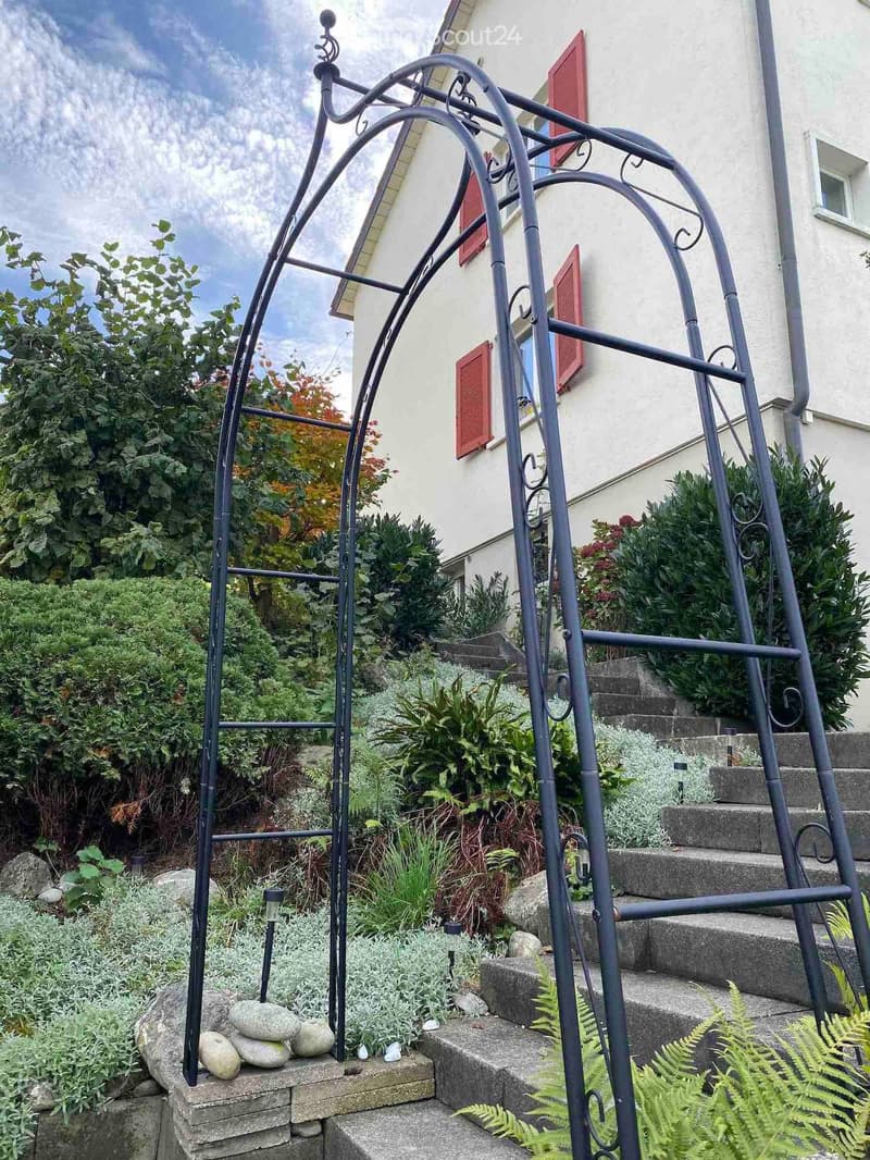 Garden area with a metal trellis, stone steps, various plants, and a small arrangement of stones on a brick platform.
