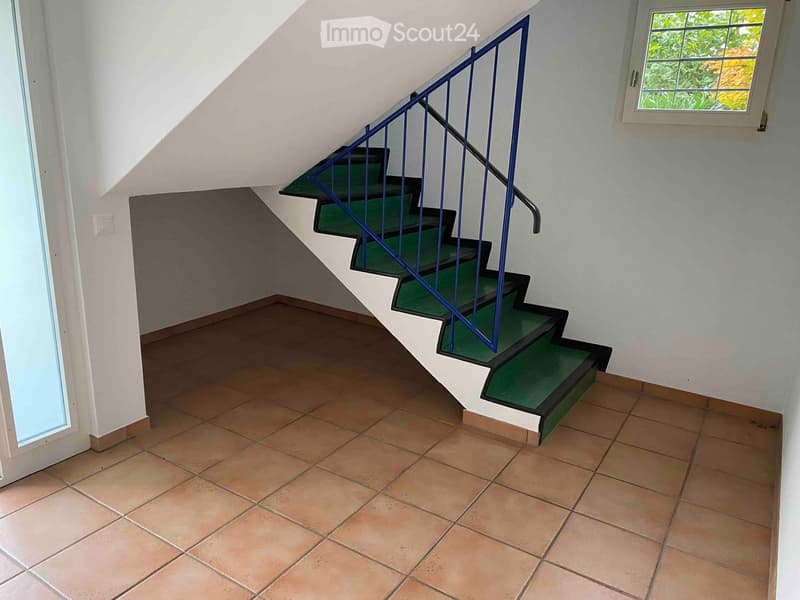 A tiled hallway with a staircase featuring green carpeted steps and a blue metal railing, leading upwards. The walls are painted white, and there are windows on either side allowing natural light to enter.