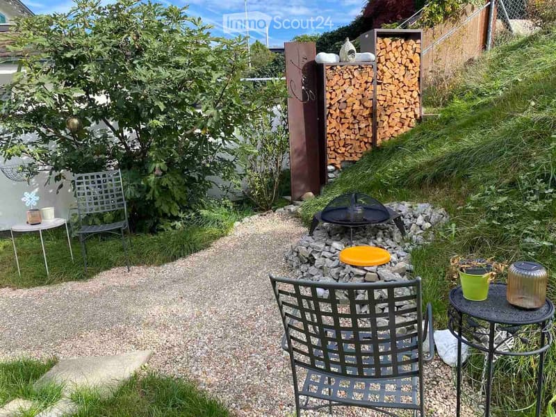 A small garden area with a gravel path, a fire pit surrounded by rocks, a stack of firewood, and outdoor furniture including chairs and tables.