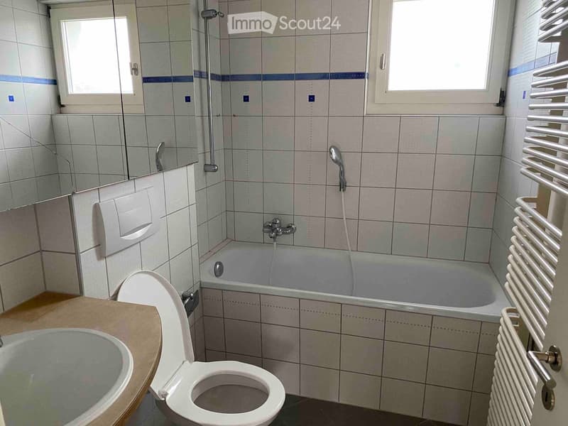 Bathroom with a toilet, bathtub, sink, radiator, and mirror. The walls are tiled with light-colored tiles and blue accents.