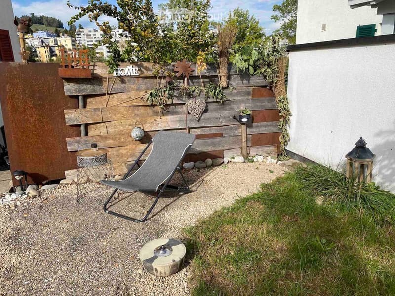 A small outdoor seating area with a wooden fence, a grey folding chair, a small round table, a lantern, and a decorative stool.