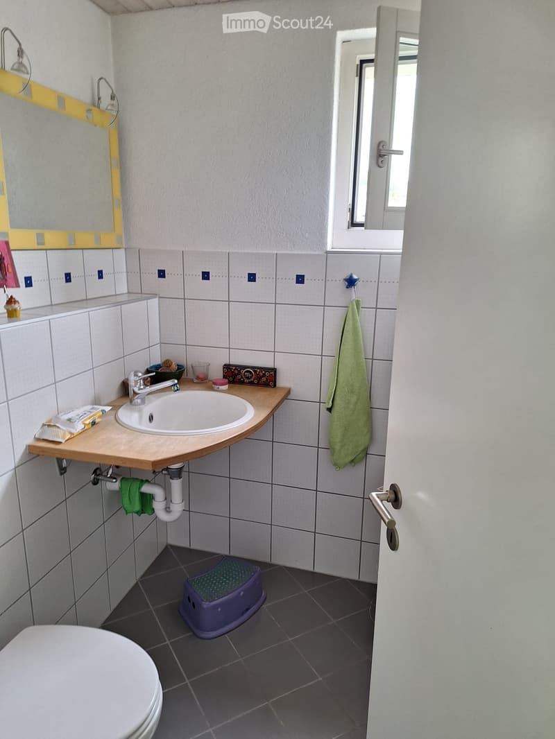 Small bathroom with white tiled walls, a wooden sink counter, a mirror, a green towel, a purple stool, and a toilet.