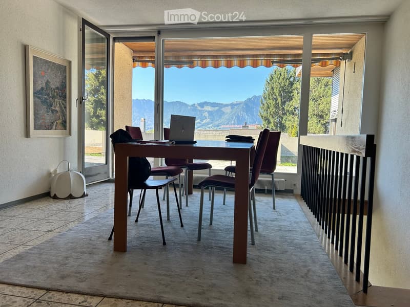 Furnished room with a table, chairs, laptop, and a view of mountains through large windows.