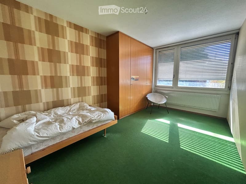 Bedroom with green carpet, wooden bed, white bedding, a white chair, and a wooden wardrobe.