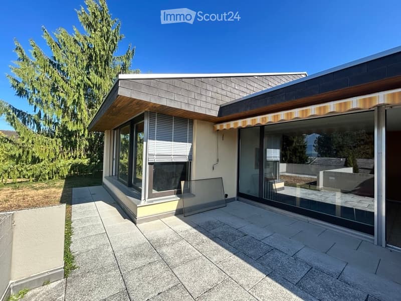 Modern two-story house with large windows, a covered patio area, and a well-maintained garden with tall trees.