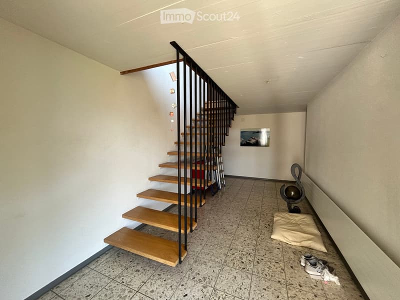 A narrow hallway with a modern staircase featuring wooden steps and metal railings, a small decorative sculpture, a cushion, and a pair of shoes on the floor.