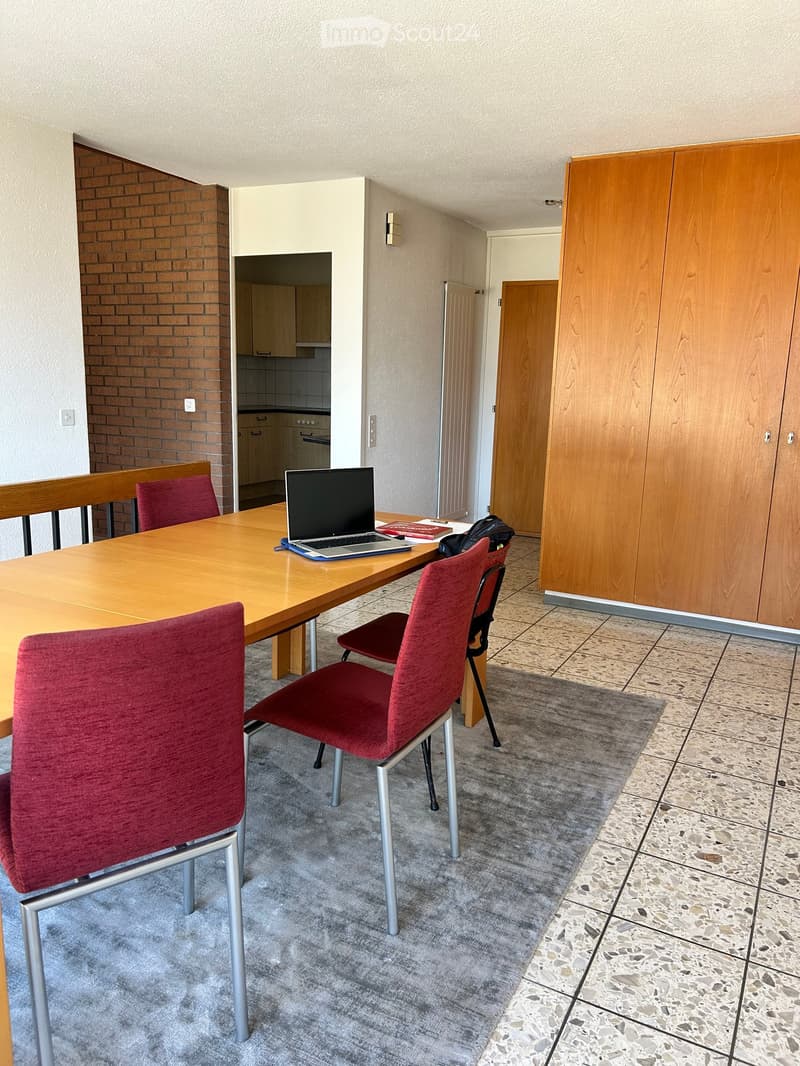 A furnished room with a wooden table, laptop, red chairs, and a wooden cupboard. The room has a tiled floor and a carpeted area.