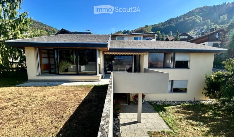 Two-story house with modern design, large windows, and a covered patio area. The house is surrounded by a well-maintained garden with a pathway leading to the entrance.